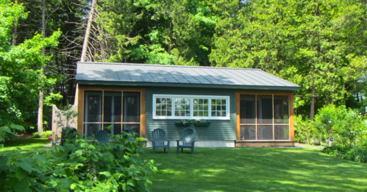 Green cottage with centered windows on a grass lawn surrounded by trees.