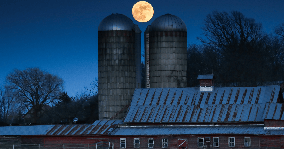 Two silos and a red Vermont barn at night with a full moon in the sky.